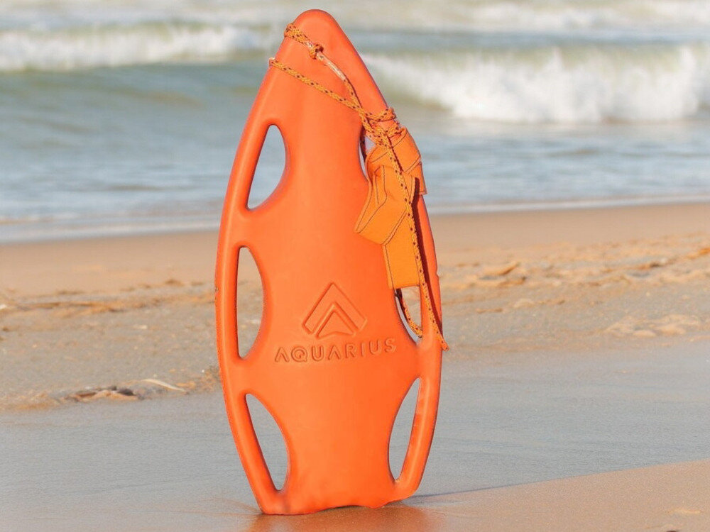 Pomarańczowa bojka ratownicza z napisem \'AQUARIUS\' stoi na piaszczystej plaży, w tle widać fale morskie.