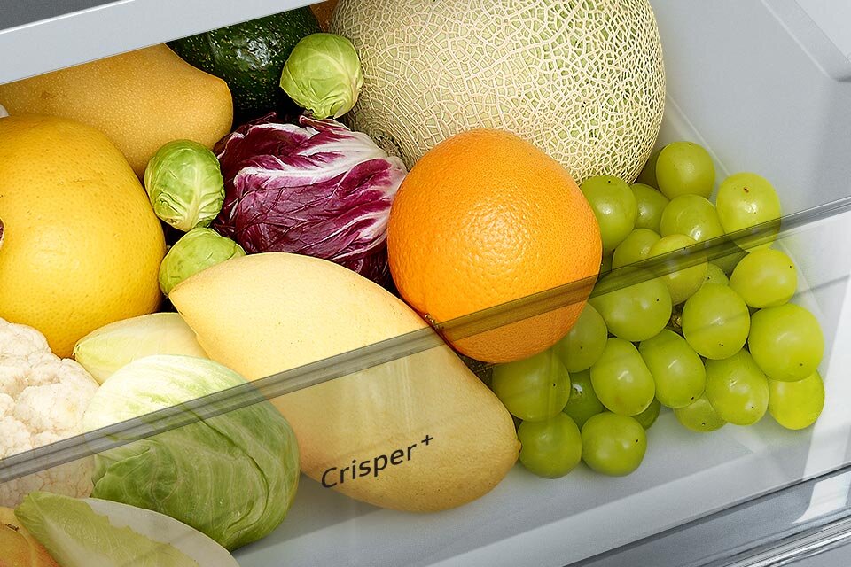Wnętrze lodówki z szufladą wypełnioną różnymi owocami i warzywami, takimi jak winogrona, pomarańcza, melon, kapusta i brukselka. Na szufladzie widoczny jest napis \'Crisper+\'.