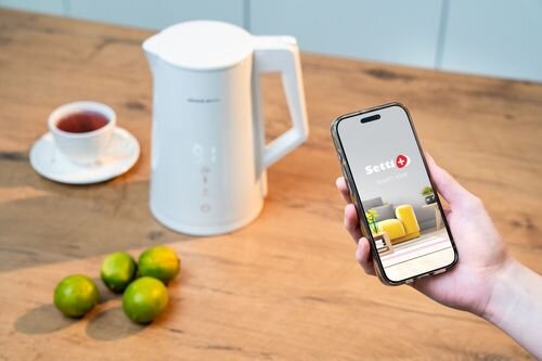 Na drewnianym blacie stoi biały czajnik elektryczny obok filiżanki herbaty i kilku limonek. Osoba trzyma smartfon z aplikacją Setti+ na ekranie.