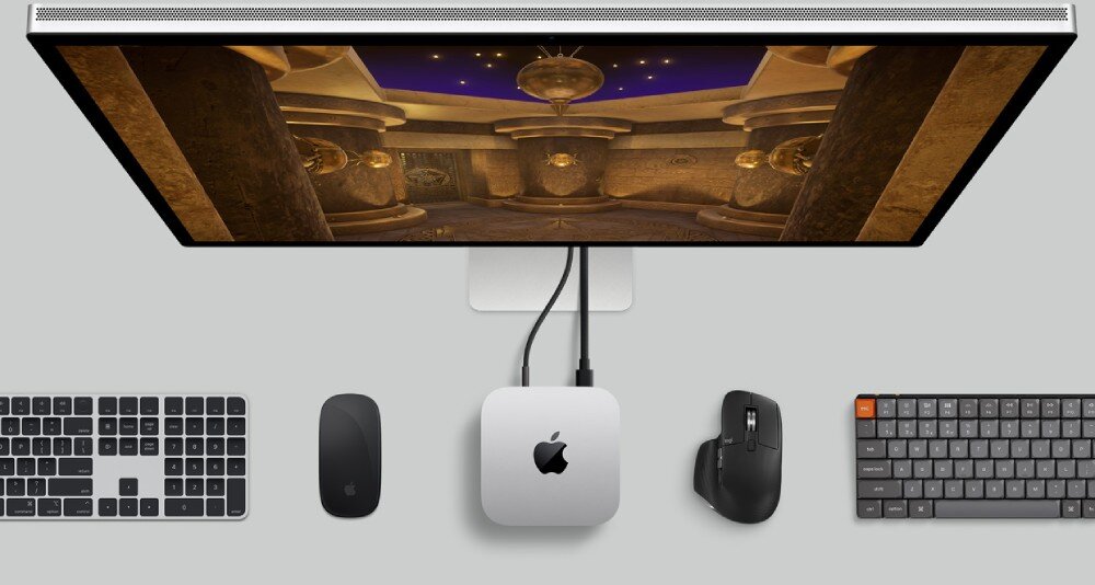 Na obrazie widoczny jest komputer Apple Mac Mini z logo Apple, podłączony do monitora, klawiatury i myszy. Na ekranie monitora wyświetlana jest gra z wnętrzem budynku.