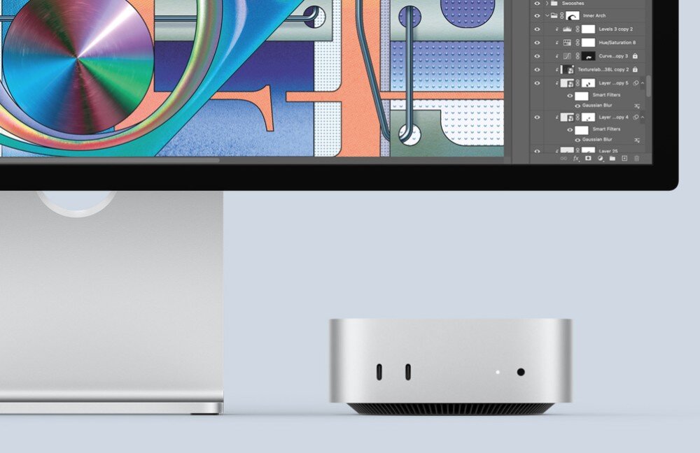Na obrazie widoczny jest srebrny komputer stacjonarny o kompaktowej budowie oraz dolna część monitora z kolorową grafiką na ekranie. Obok komputera znajduje się logo APPLE.