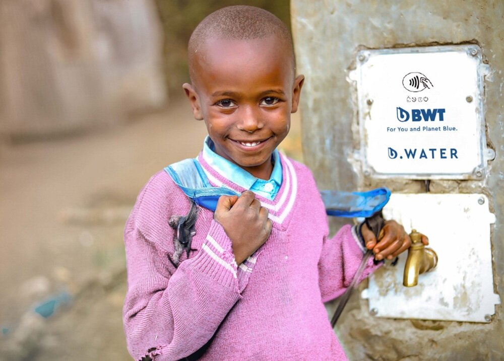 Dziecko w różowym swetrze stoi obok kranu z wodą. Na ścianie obok kranu widnieje logo BWT z napisem \'For You and Planet Blue. B.WATER\'.