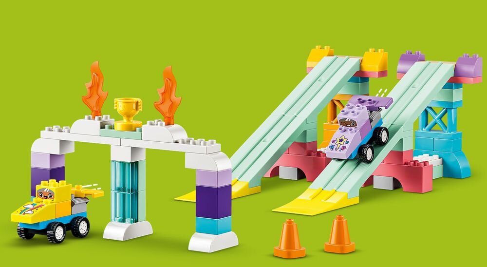 Na zielonym tle widoczne są dwa zestawy torów z klocków LEGO, z pojazdami i dekoracjami w postaci pucharów i płomieni. Obok torów znajdują się dwa pomarańczowe pachołki.