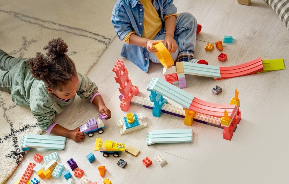 Dwoje dzieci bawi się zestawem klocków, tworząc rampy i pojazdy z kolorowych elementów. Na podłodze widać różnorodne klocki LEGO w jasnych kolorach.