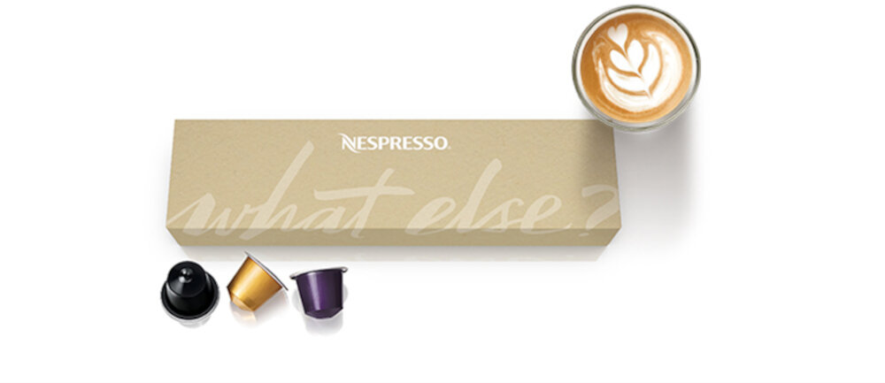 Na obrazie znajduje się pudełko z napisem \'NESPRESSO what else?\' oraz trzy kapsułki do kawy w różnych kolorach. Obok pudełka widoczna jest filiżanka kawy z mleczną pianką na wierzchu.