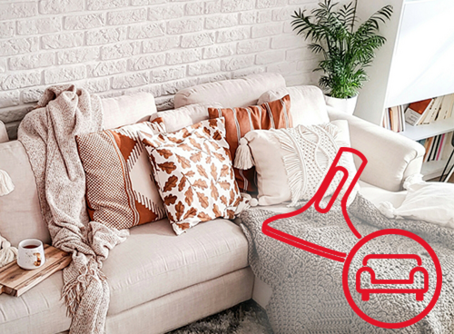 Jasna sofa z kilkoma poduszkami w różnych wzorach i kolorach, przykryta kocem. Na obrazie widoczny jest czerwony symbol odkurzacza z ikoną sofy.