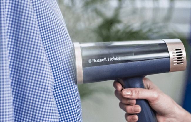 Osoba używa parownicy do ubrań marki Russell Hobbs na koszuli w kratkę. Na urządzeniu widoczne jest logo \'Russell Hobbs\'.