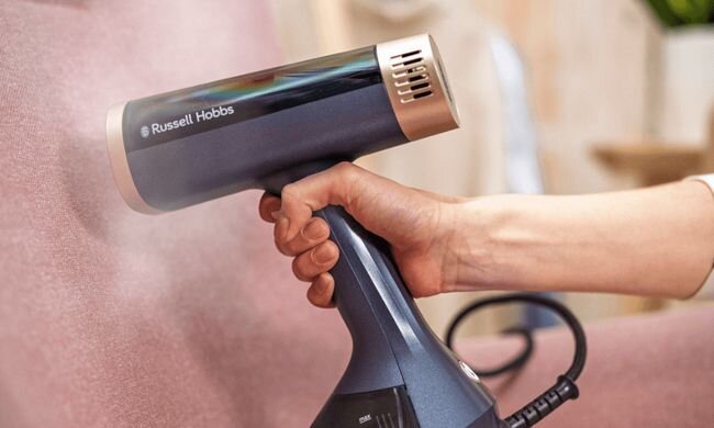 Osoba trzyma parownicę do ubrań, z której wydobywa się para. Na urządzeniu widoczne jest logo \'Russell Hobbs\'.
