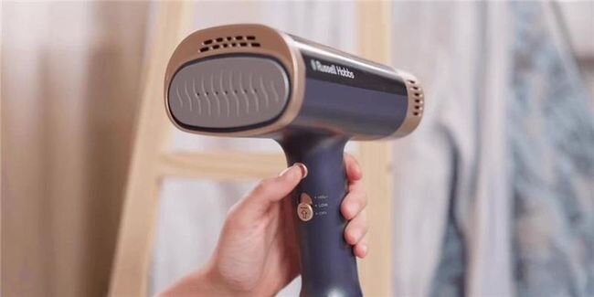 Osoba trzyma parownicę do ubrań w kolorze granatowym z elementami złotymi. Na urządzeniu widoczne jest logo \'Russell Hobbs\'.