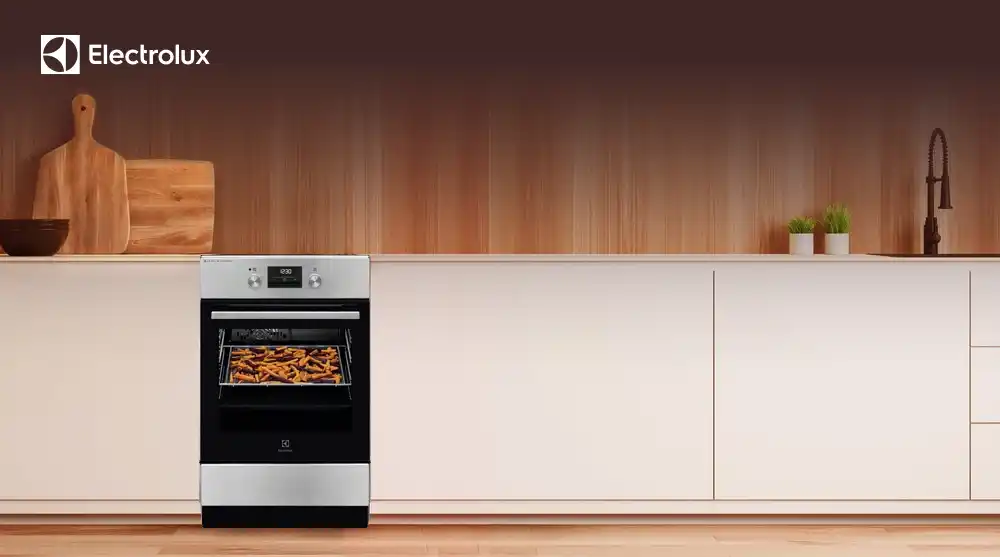Na obrazie znajduje się kuchnia z piekarnikiem Electrolux, w którym pieką się frytki. W tle widoczne są drewniane panele, deska do krojenia i zlew z roślinami w doniczkach.