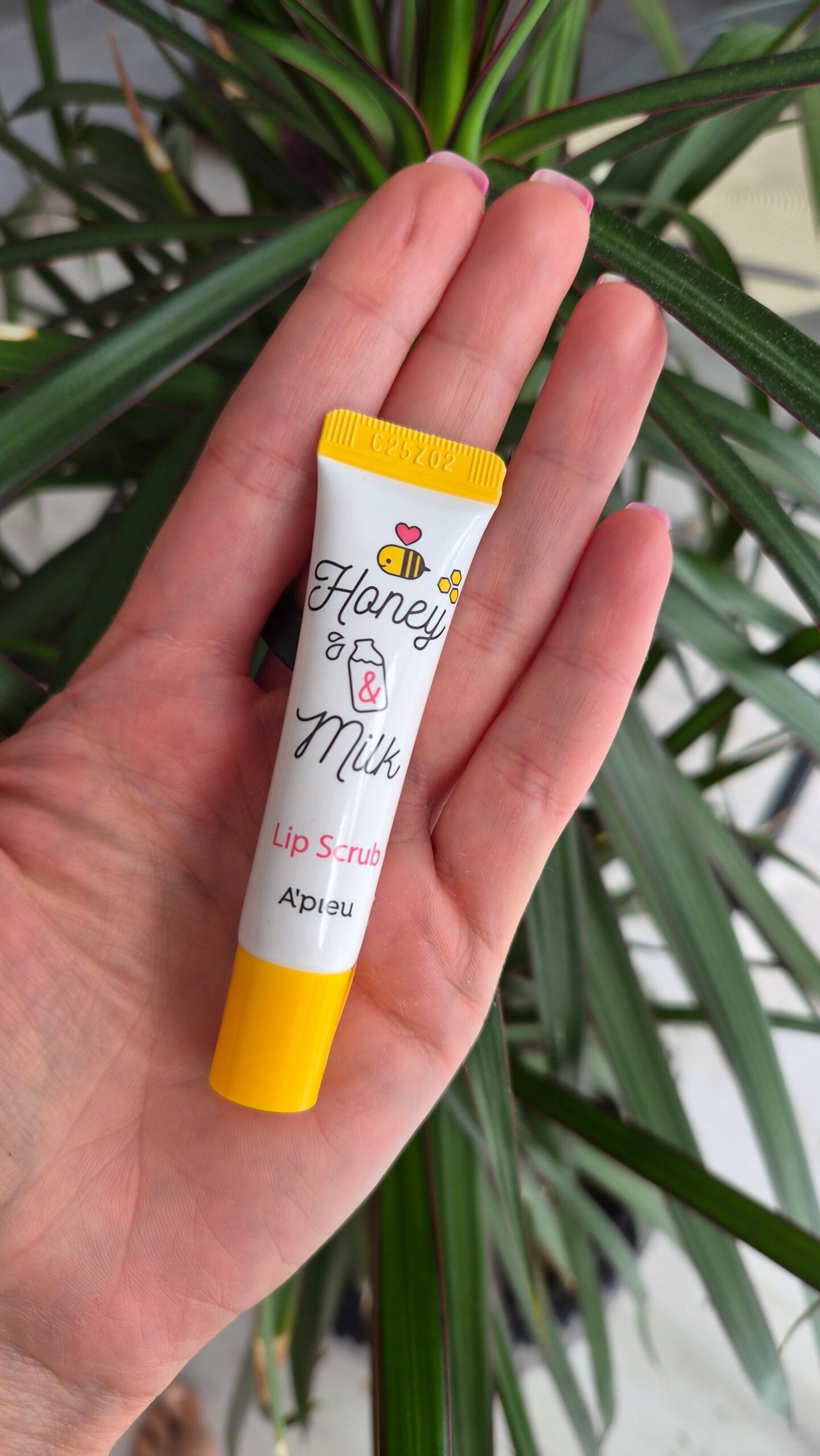 Osoba trzyma w dłoni tubkę z napisem \'Honey & Milk Lip Scrub A\'PIEU\'. W tle widoczne są zielone liście rośliny.