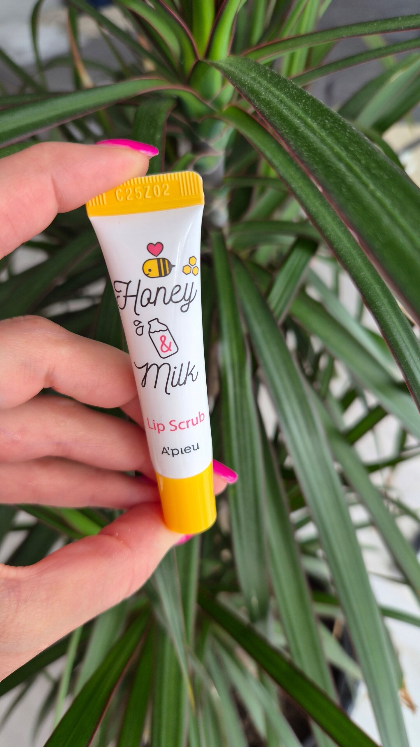 Osoba trzyma tubkę produktu z napisem \'Honey & Milk Lip Scrub A\'Pieu\' na tle zielonej rośliny. Tubka jest biała z żółtą zakrętką i ozdobiona grafiką pszczoły oraz kwiatka.