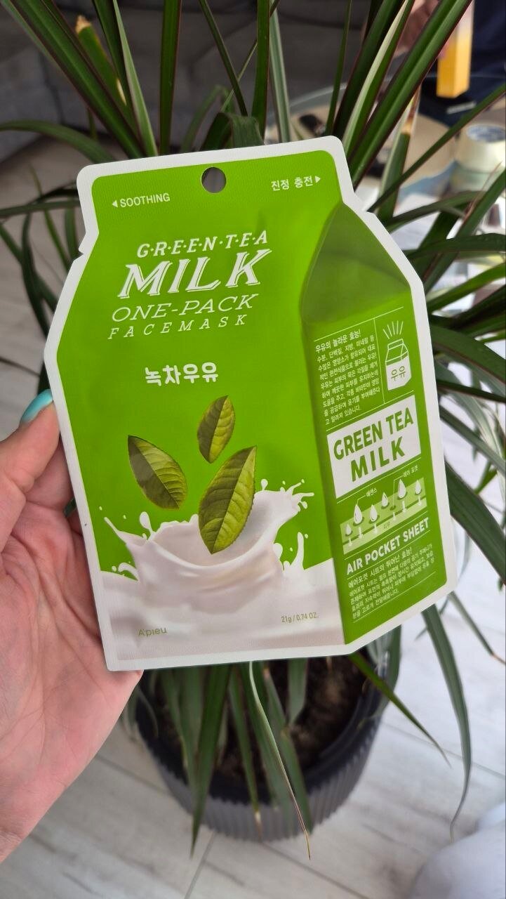 Opakowanie maseczki w płachcie z zieloną herbatą, z napisem \'GREEN TEA MILK ONE-PACK FACE MASK\' oraz grafiką liści herbaty w mleku. W tle widoczne są zielone liście rośliny doniczkowej.