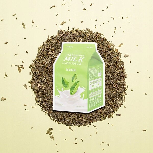 Opakowanie maseczki w płachcie A\'PIEU Green Tea Milk One-Pack 21 g leży na stosie suszonych liści herbaty. Na opakowaniu widoczny jest napis \'GREEN TEA MILK ONE-PACK\' oraz grafika przedstawiająca liście herbaty i mleko.