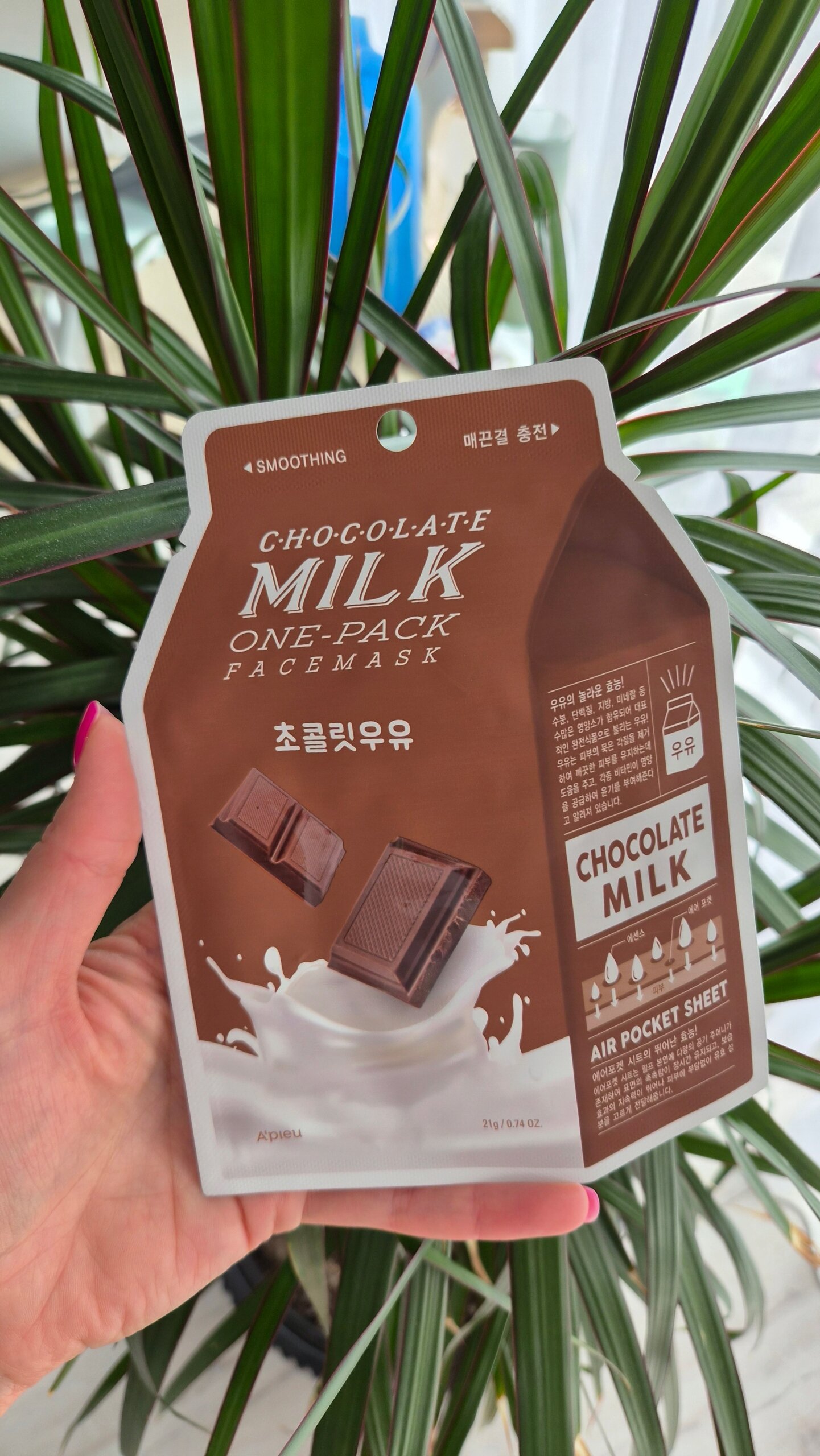 Opakowanie maseczki w płachcie o nazwie \'Chocolate Milk One-Pack Face Mask\' z grafiką kostek czekolady i mleka. W tle widoczne są zielone liście rośliny.