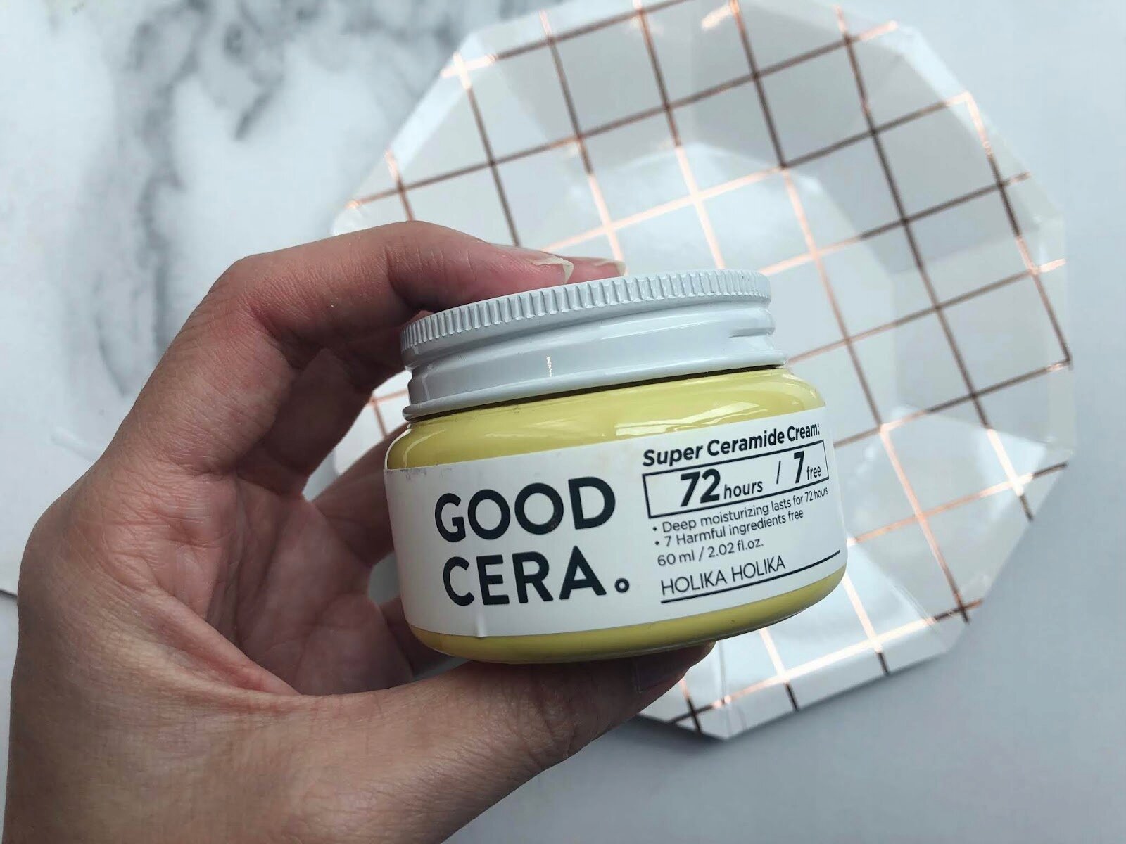 Osoba trzyma żółty słoik z kremem do twarzy, na którym widnieje napis \'GOOD CERA. Super Ceramide Cream 72 hours 7 free Deep moisturizing lasts for 72 hours 7 Harmful ingredients free 60 ml / 2.02 fl.oz. HOLIKA HOLIKA\'. W tle znajduje się talerz z geometrycznym wzorem.
