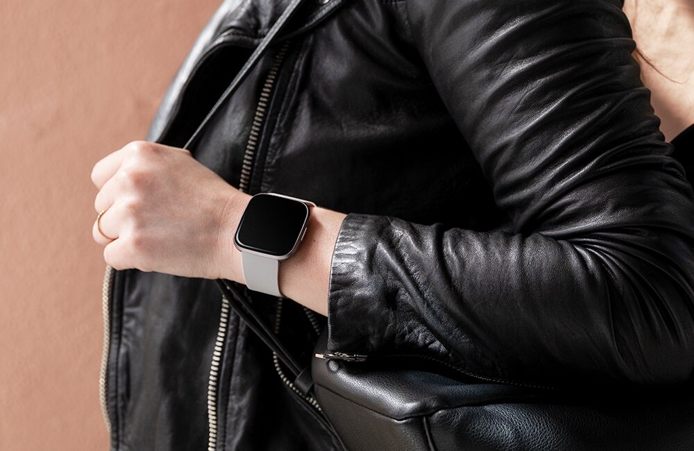 Osoba w czarnej skórzanej kurtce nosi szary smartwatch na nadgarstku. Tło jest w kolorze jasnobrązowym.