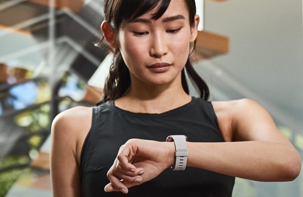 Osoba w czarnej koszulce patrzy na szary smartwatch na nadgarstku. W tle widoczne są rozmyte elementy wnętrza budynku.
