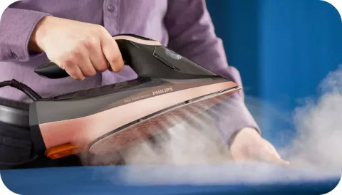 Osoba prasuje niebieski materiał przy użyciu żelazka, z którego wydobywa się para. Na żelazku widoczny jest napis \'PHILIPS\'.