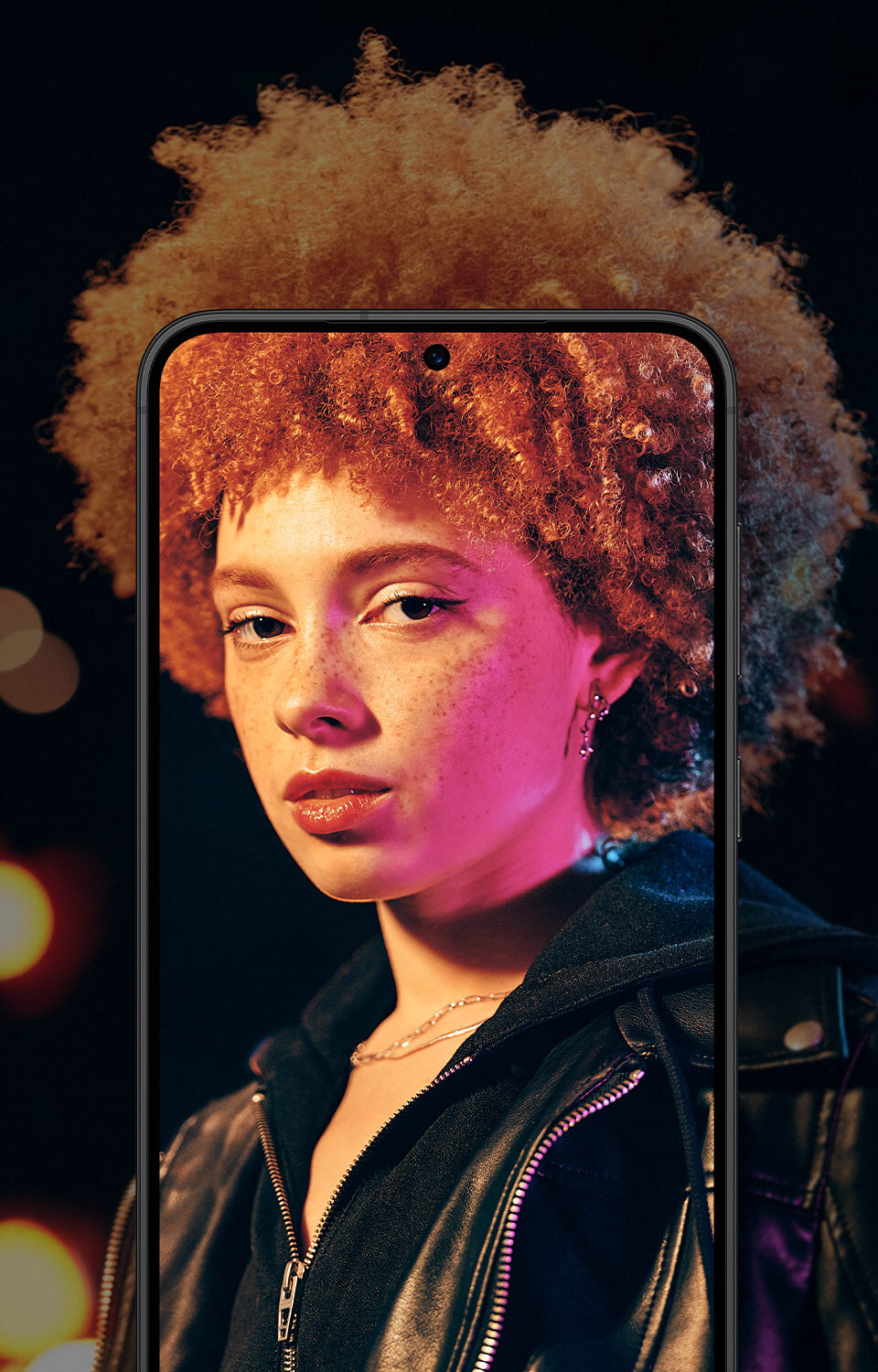 Smartfon z wyświetlaczem pokazującym osobę z kręconymi włosami w ciemnym otoczeniu z kolorowymi światłami w tle.
