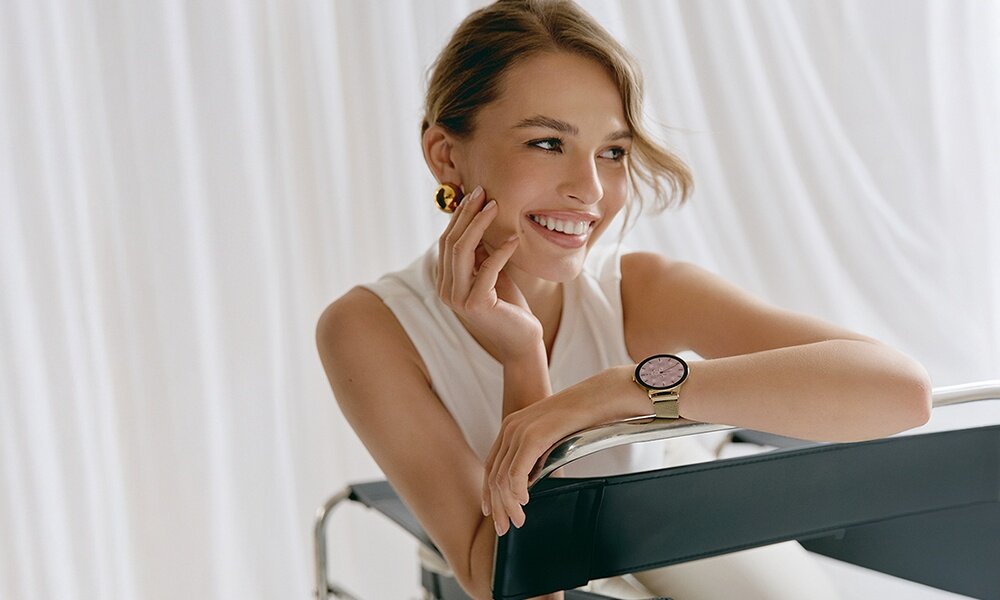 Osoba siedzi przy metalowej konstrukcji, nosząc złoty smartwatch z okrągłą tarczą na nadgarstku. W tle widoczne są białe zasłony.