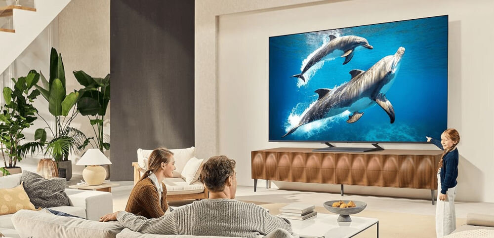 W salonie na ścianie wisi duży telewizor, na którym wyświetlany jest obraz delfinów pływających w wodzie. W pomieszczeniu znajdują się trzy osoby siedzące na kanapie oraz dziecko stojące obok telewizora.