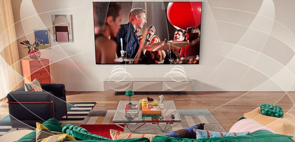W przestronnym salonie na ścianie wisi duży telewizor, na którym wyświetlany jest obraz koncertu. W pomieszczeniu znajdują się kolorowe meble i dekoracje, a na ekranie widoczne są efekty dźwiękowe.
