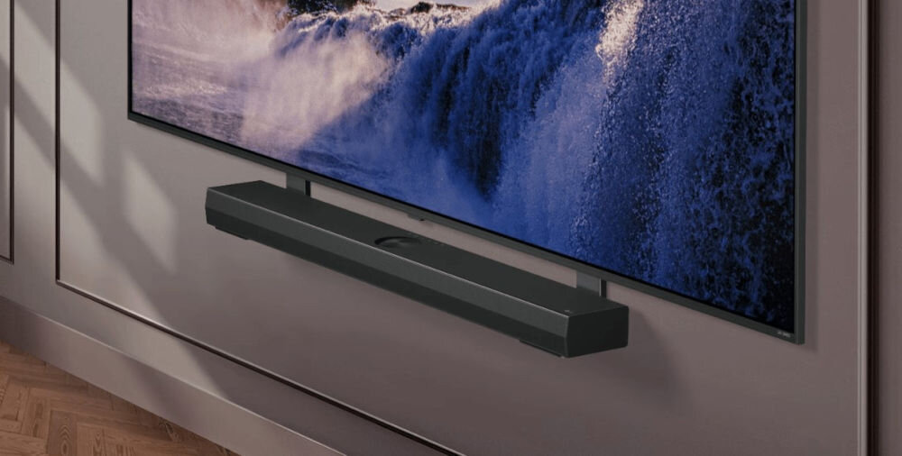 Na obrazie widoczny jest telewizor z wyświetlanym obrazem fal morskich oraz zamontowany poniżej soundbar. Telewizor jest umieszczony na ścianie w pomieszczeniu z drewnianą podłogą.