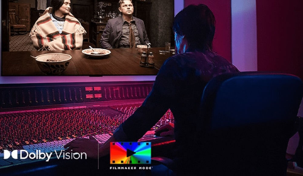 Osoba siedzi przed dużym panelem sterującym, patrząc na ekran, na którym widać dwie osoby przy stole. Na dole obrazu znajdują się logotypy \'Dolby Vision\' i \'Filmmaker Mode\'.