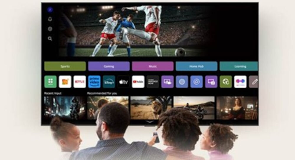 Na ekranie telewizora wyświetlany jest mecz piłki nożnej oraz interfejs z aplikacjami takimi jak Netflix, Prime Video i YouTube. Przed telewizorem siedzą cztery osoby, patrząc na ekran.