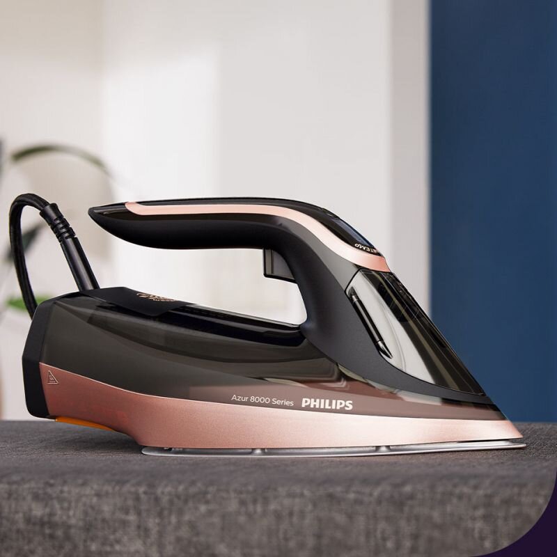 Żelazko w kolorze czarno-różowym z widocznym napisem \'Azur 8000 Series PHILIPS\' stoi na szarej powierzchni. W tle znajduje się rozmyte wnętrze pomieszczenia.