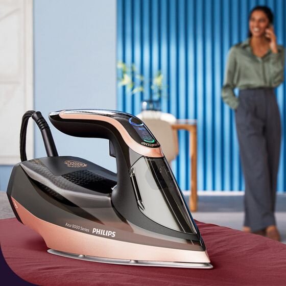 Żelazko PHILIPS Azur 8000 Series DST8041/80 stoi na czerwonym materiale. W tle widoczna jest osoba oraz niebieska ściana z pionowymi żaluzjami.