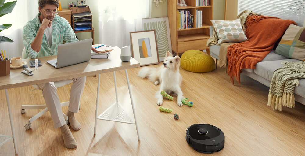 W jasnym pokoju z drewnianą podłogą znajduje się robot sprzątający oraz pies leżący obok zabawek. Przy biurku siedzi osoba pracująca na laptopie, a w tle widać sofę z poduszkami i kocem.