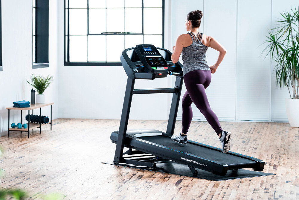 Osoba biegnie na bieżni elektrycznej w jasnym pomieszczeniu z drewnianą podłogą. Na bieżni widoczny jest napis \'HORIZON FITNESS\'.