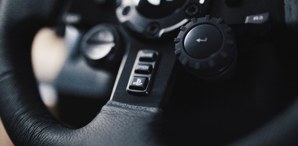 Zbliżenie na fragment kierownicy z przyciskami, w tym jeden z logo PlayStation. Widoczna jest także tekstura czarnej skóry na kierownicy.