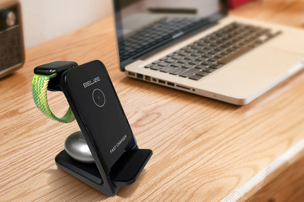 Na drewnianym biurku znajduje się czarna ładowarka indukcyjna z napisem \'BELINE\' i \'FAST CHARGER\', obok której leży otwarty laptop. Na ładowarce umieszczony jest smartwatch z zielonym paskiem.