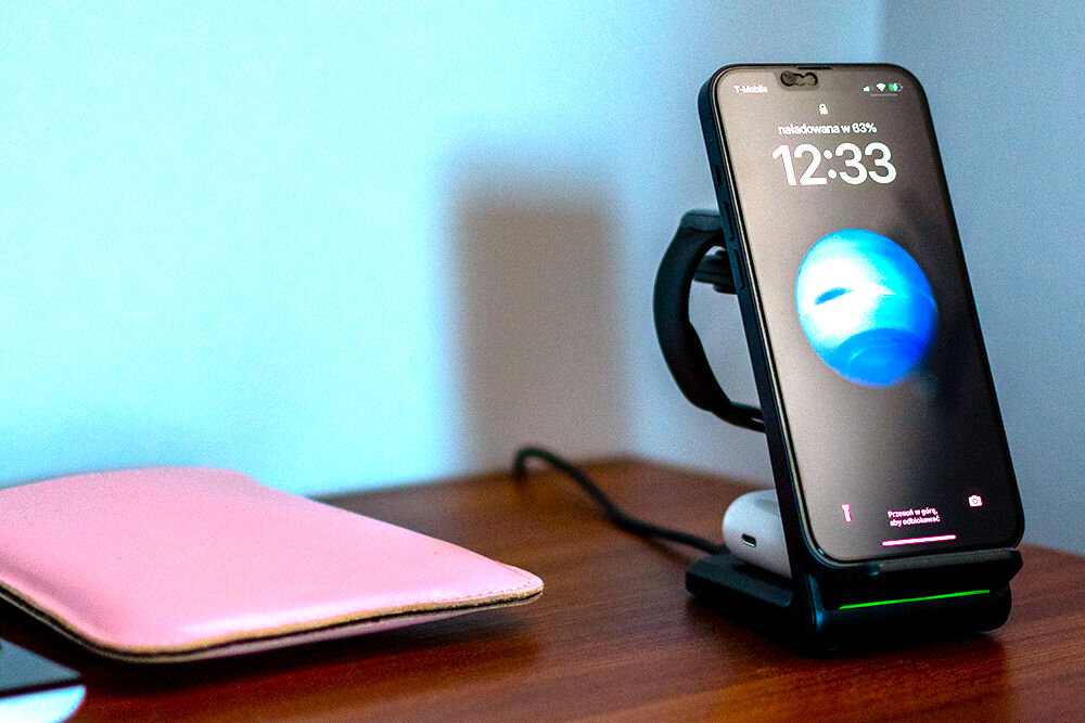 Na drewnianym blacie stoi telefon na czarnej ładowarce indukcyjnej, z wyświetlaczem pokazującym godzinę 12:33 i napis \'naładowana w 63%\'. Obok leży różowy futerał.