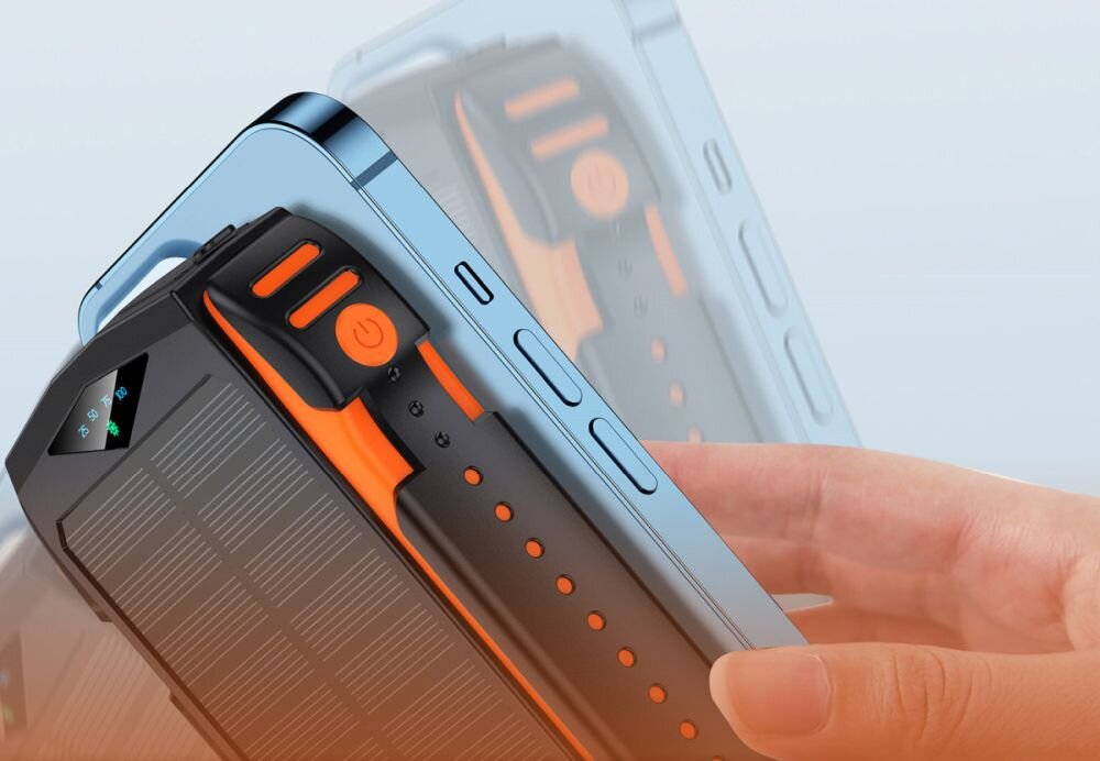 Na zdjęciu widać czarny powerbank z pomarańczowymi elementami, trzymany w dłoni obok smartfona. Powerbank ma widoczny przycisk zasilania i wskaźnik LED.