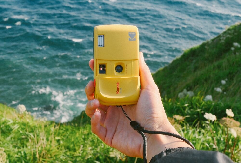 Osoba trzyma żółty aparat analogowy z logo Kodak, na tle morza i zielonego klifu.