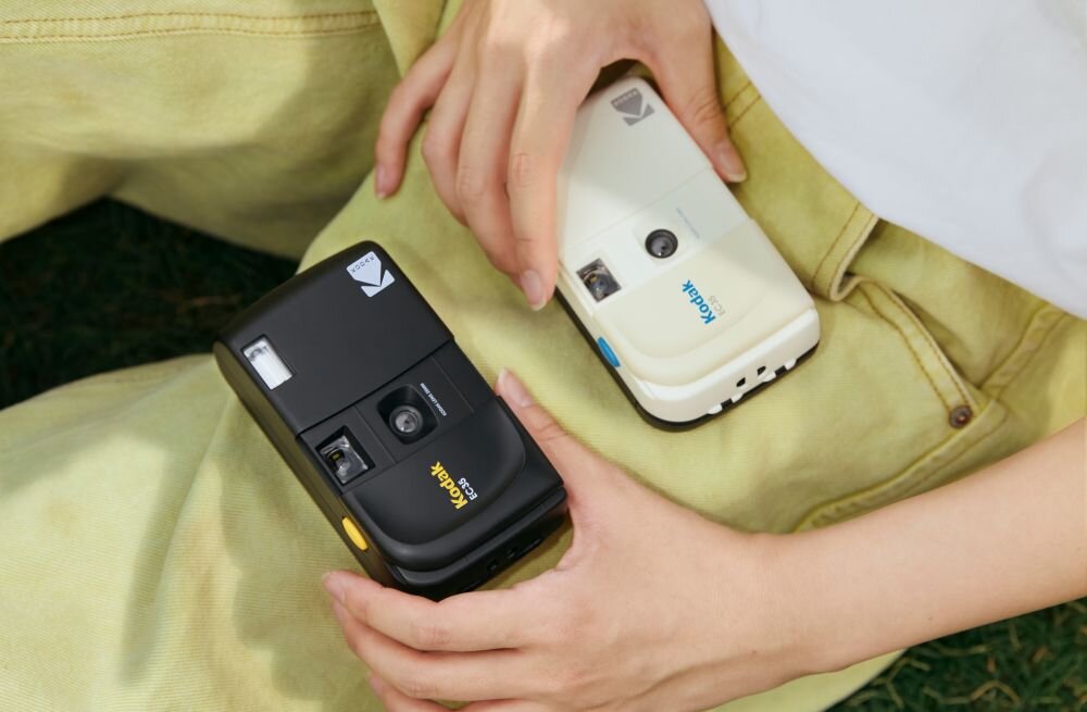 Osoba trzyma dwa aparaty analogowe marki Kodak, jeden czarny z żółtym napisem \'Kodak\' i drugi biały z niebieskim napisem \'Kodak\'.