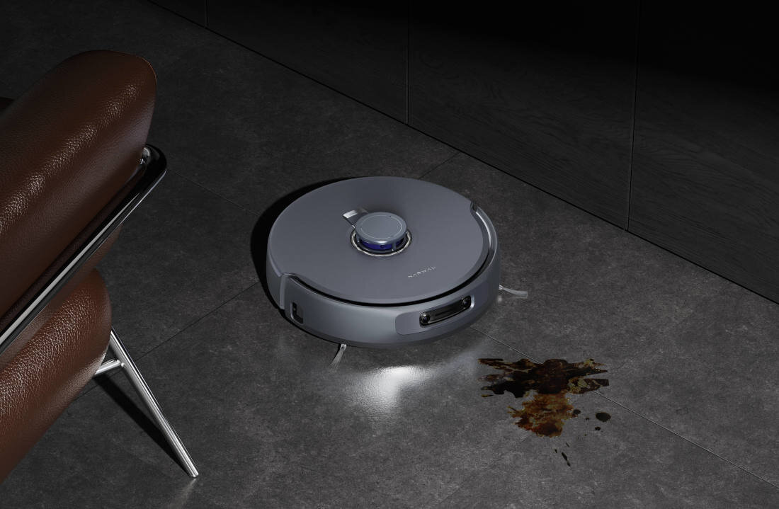 Na ciemnej podłodze znajduje się szary robot sprzątający z widocznym logo \'NARWAL\'. Obok robota widoczna jest plama na podłodze.
