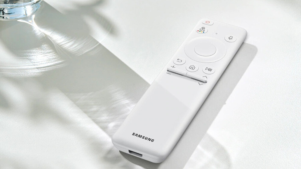 Na obrazie widoczny jest biały pilot z logo \'SAMSUNG\' na dolnej części. Pilot leży na jasnej powierzchni, a obok niego widać cień i odbicie światła.