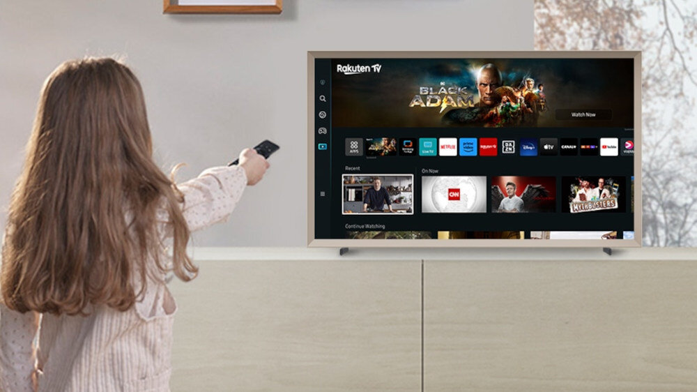 Osoba trzymająca pilot skierowany w stronę telewizora, na którym wyświetlone jest menu z aplikacjami takimi jak Netflix, YouTube i Rakuten TV. Na ekranie widoczny jest także film \'Black Adam\'.
