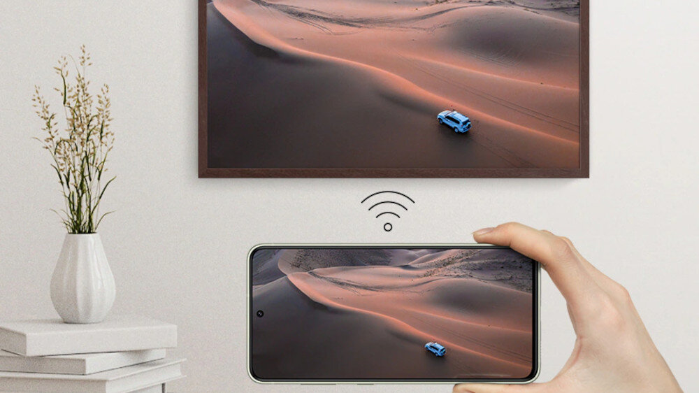 Na ścianie wisi telewizor z wyświetlanym obrazem pustyni z niebieskim samochodem. Przed telewizorem znajduje się ręka trzymająca smartfon, na którym wyświetlany jest ten sam obraz, a pomiędzy nimi widoczna jest ikona sygnału bezprzewodowego.