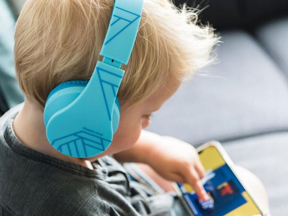 Dziecko nosi niebieskie słuchawki z geometrycznym wzorem, patrząc na ekran urządzenia mobilnego. Na słuchawkach widoczny jest napis \'PowerLocus\'.