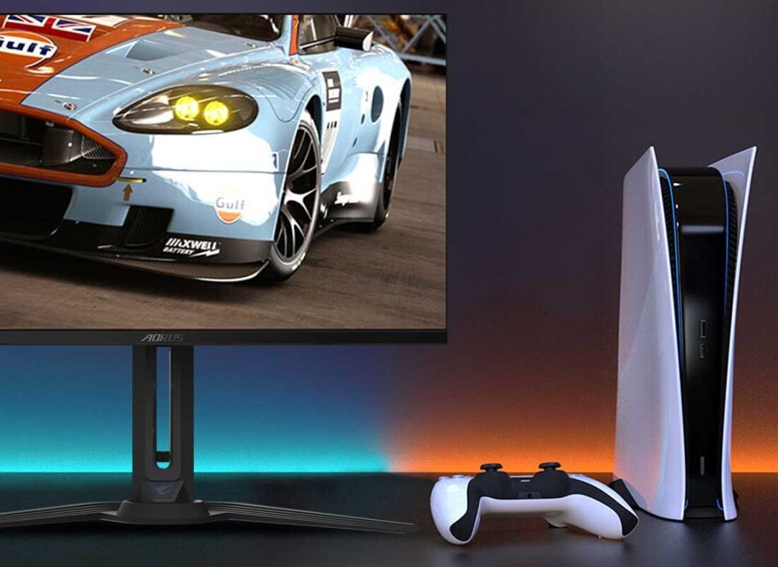 Na obrazie widoczny jest monitor z wyświetlonym samochodem wyścigowym oraz konsola do gier z kontrolerem. Na monitorze znajduje się logo \'AORUS\'.