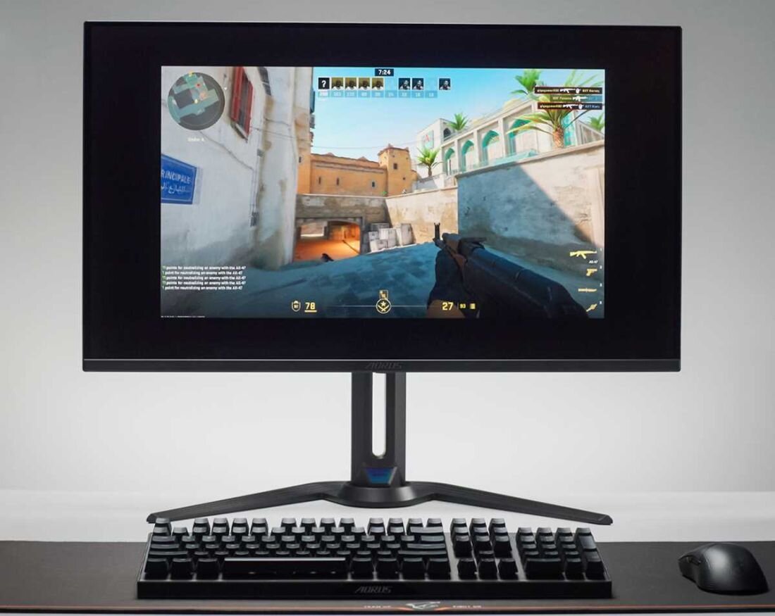 Monitor wyświetla grę FPS z widocznym interfejsem użytkownika i postacią trzymającą broń. Na biurku przed monitorem znajduje się klawiatura i myszka.