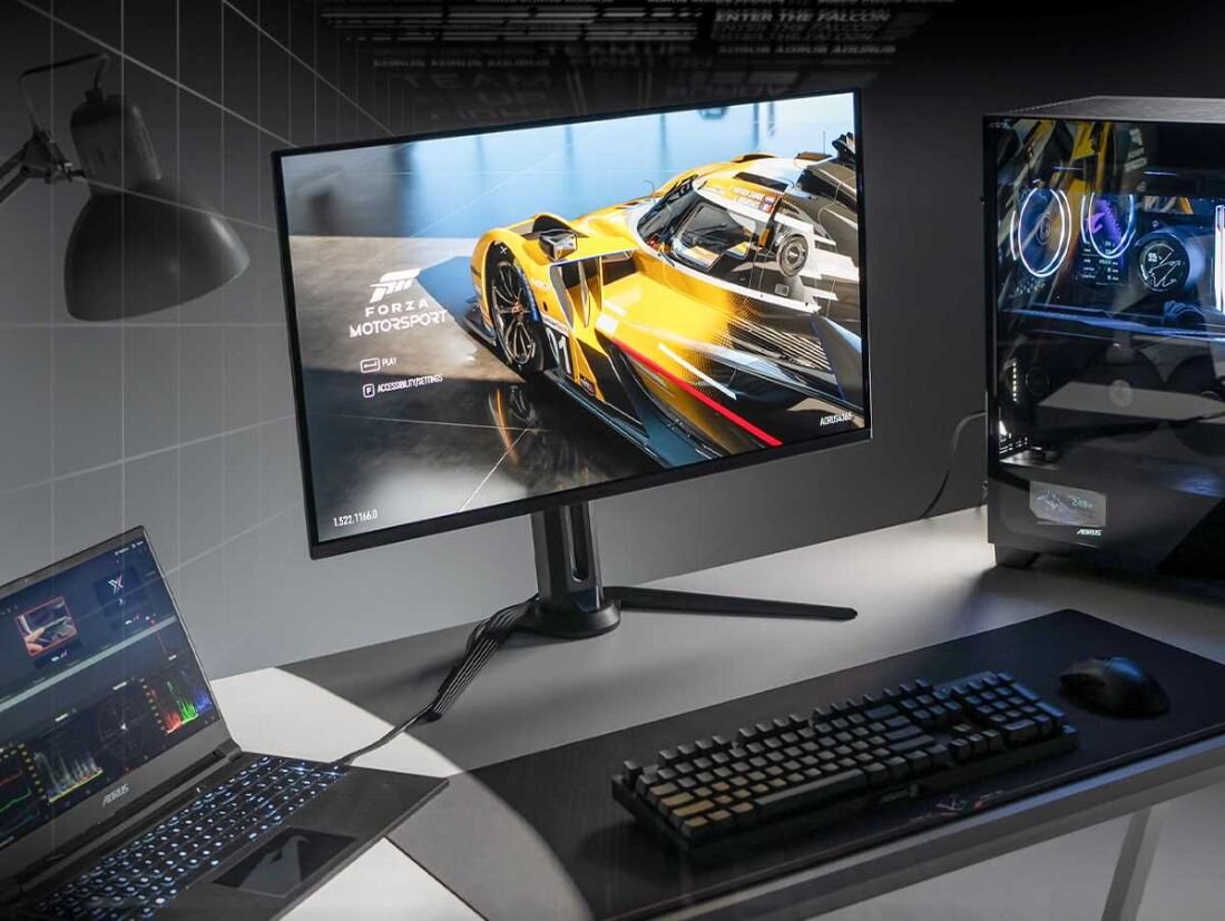 Na biurku znajduje się monitor wyświetlający grę wyścigową z żółtym samochodem, obok laptop i komputer stacjonarny z podświetleniem RGB. Na ekranie monitora widoczny jest tekst: \'Forza Motorsport\'.