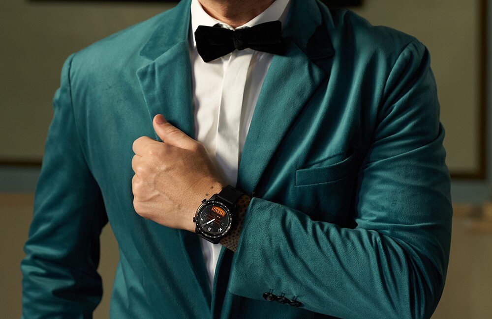 Osoba w eleganckim, zielonym garniturze z muszką, nosząca czarny smartwatch na nadgarstku.