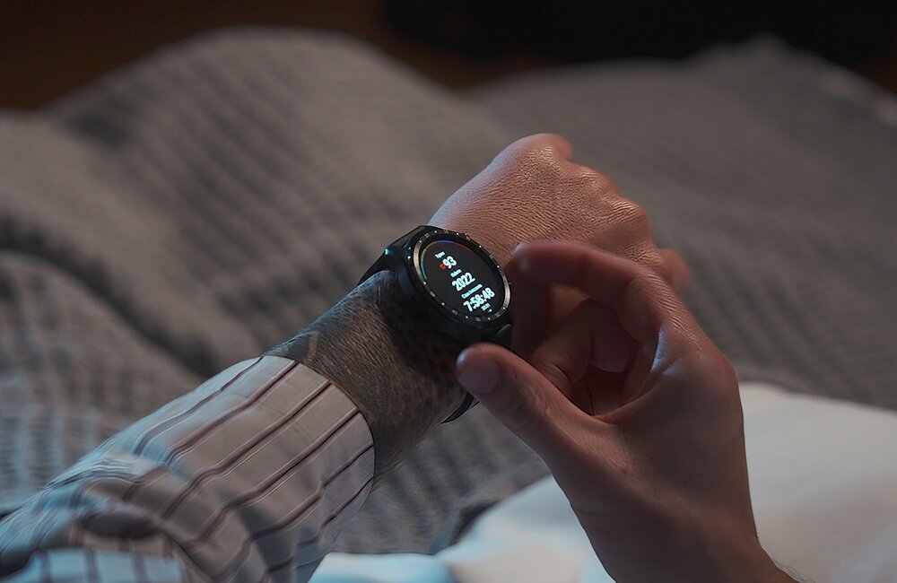 Osoba nosi czarny smartwatch na nadgarstku, na ekranie widoczne są dane dotyczące zdrowia i czasu. Tło jest rozmyte, a osoba ma na sobie koszulę w paski.